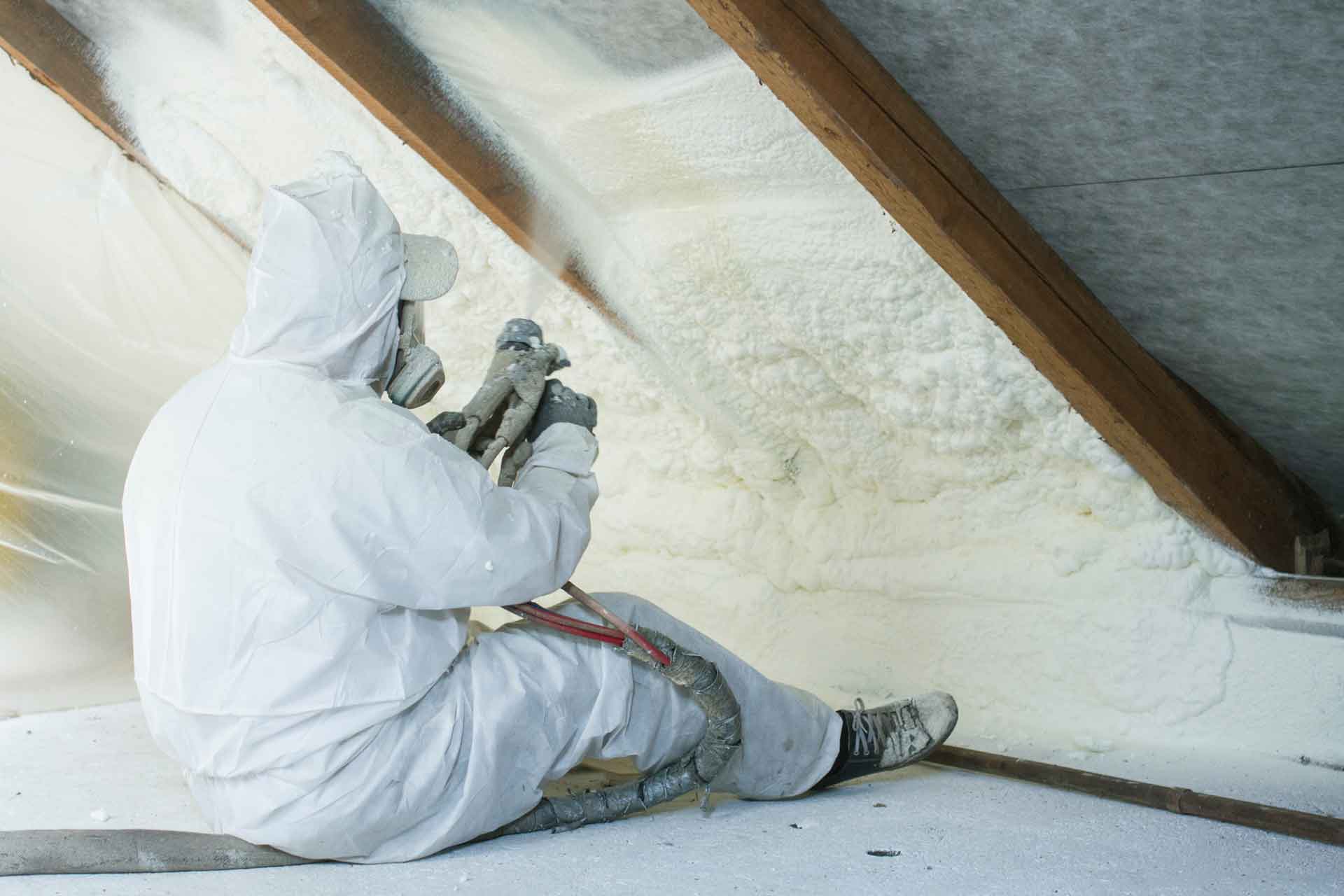 Man sitting on floor in attic while he sprays foam insulation on the ceiling